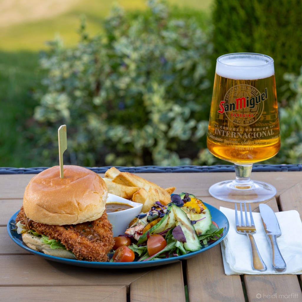 A fried chicken burger served at the table on a plate, next to a pint of San Miguel lager. 