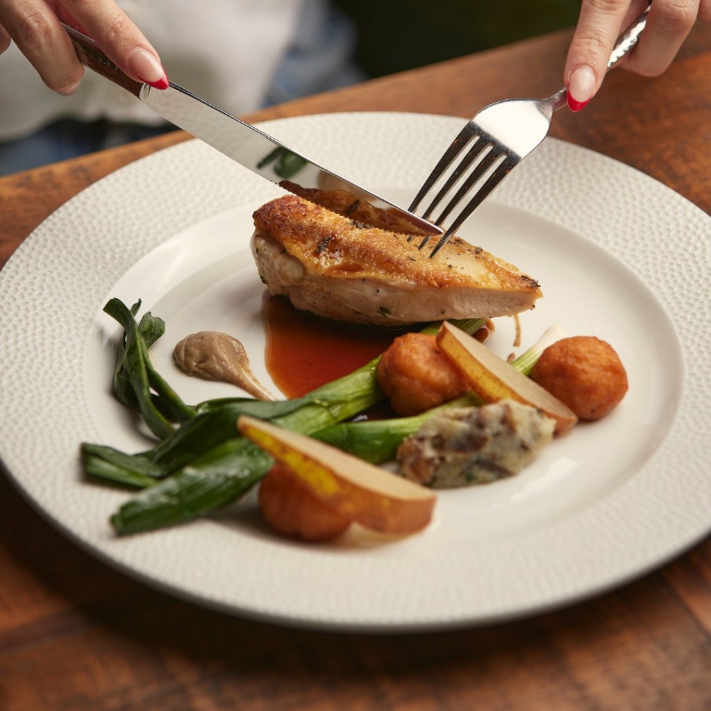 A close-up of a fine dining chicken dish, as someone holds their knife and fork above the meat, ready to cook. 